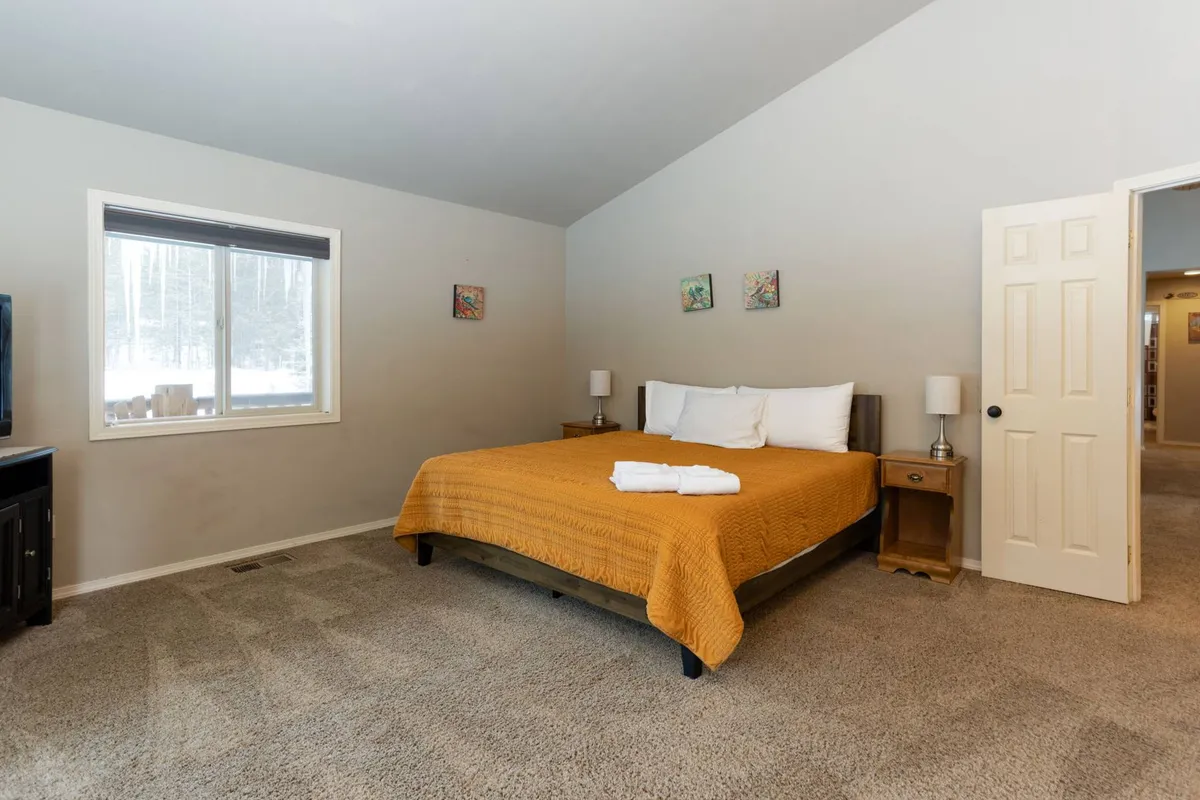 A cozy bedroom featuring a bed with an orange quilt, a large window offering a view of the outdoors, a nightstand with a lamp, and decorative artwork. Natural light streams in, suggesting a connection to the surrounding landscape and outdoor adventure possibilities.