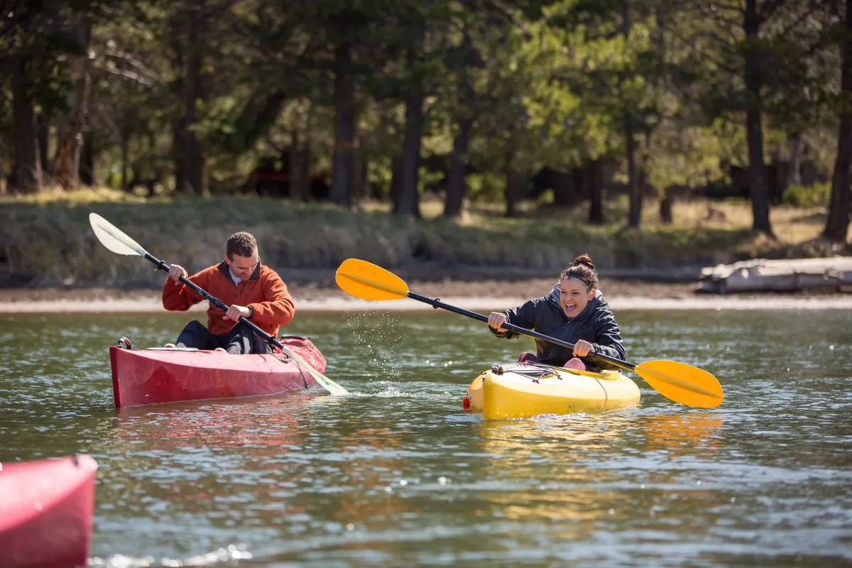 Two kayakers paddle on calm waters surrounded by lush, green banks. The peaceful yet playful scene reflects summer outdoor activities and family adventure, set in beautiful Central Oregon nature.