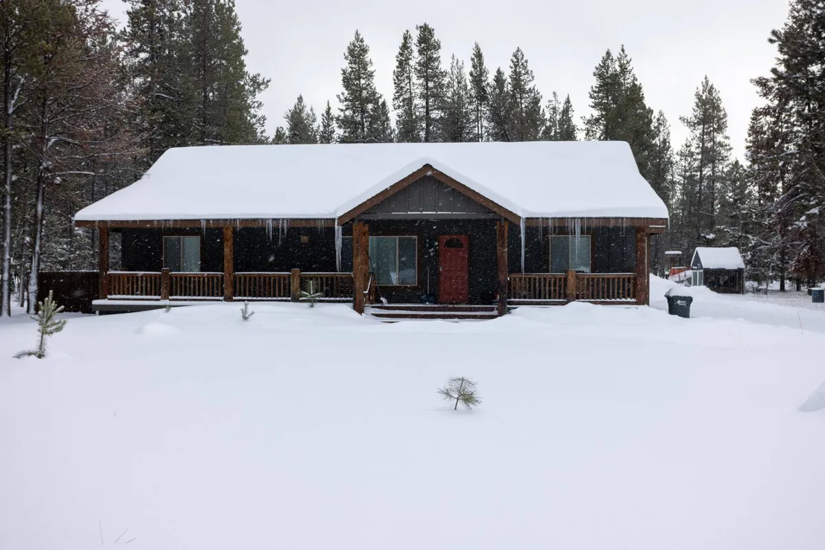 A welcoming, snow-covered house with a wooden porch and bright red door, surrounded by fresh winter snow. The serene scene suggests cozy moments spent watching snowfall from the porch, building snowmen, or returning inside to warm up after outdoor adventures.