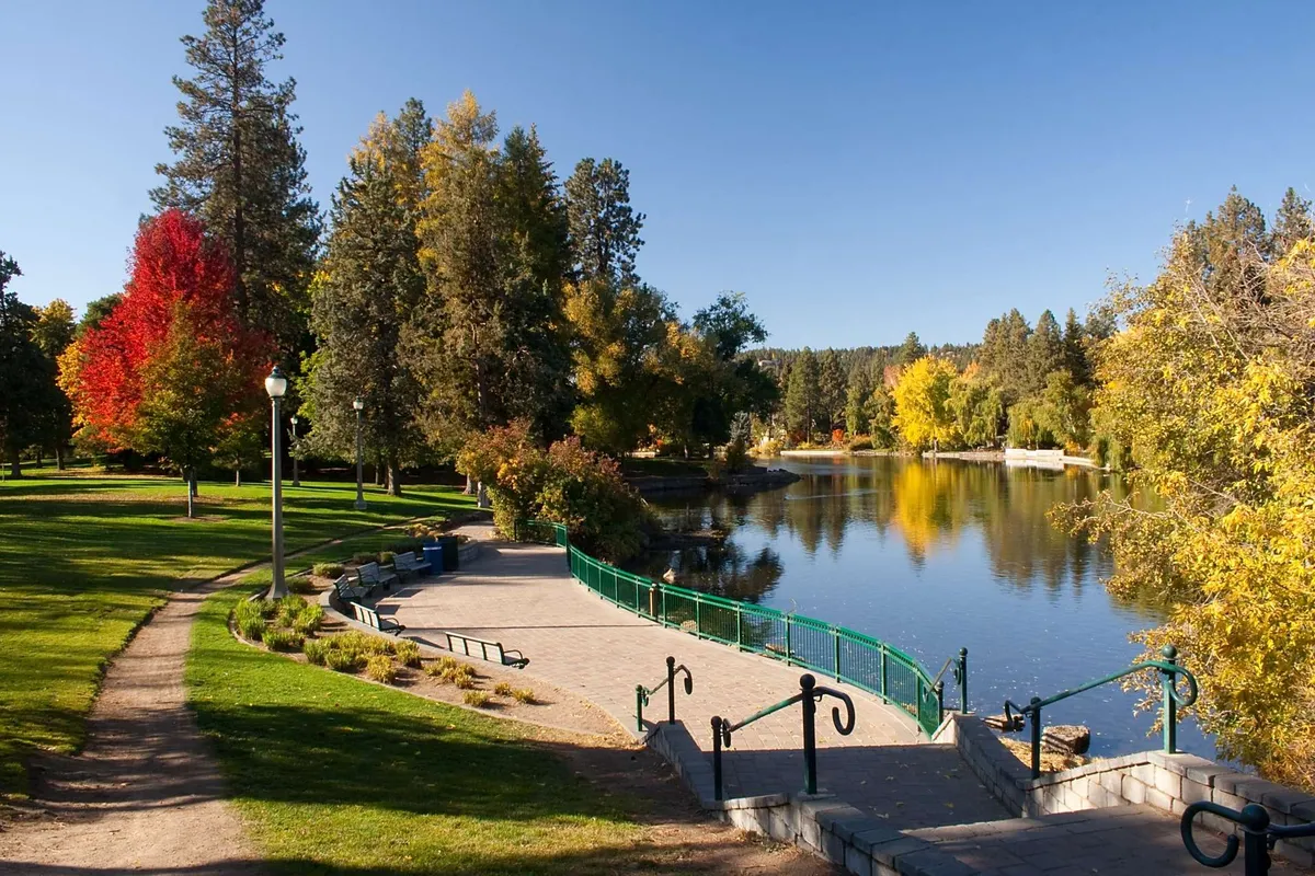 An exterior scene featuring a serene lake surrounded by trees, a pathway suitable for walks, benches for resting, and a small bridge. The setting is tranquil and clearly designed for outdoor enjoyment and strolls.