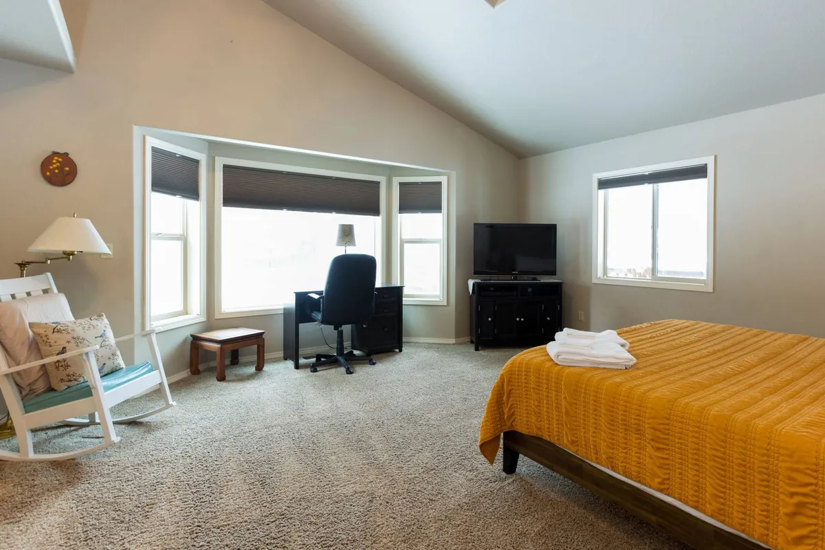 This image highlights a spacious bedroom featuring a yellow bedspread, a cozy rocking chair, a desk and chair, and large windows that flood the room with natural light. The overall ambiance is tranquil and comfortable, suggesting a restful space ideal for recharging or getting some work done.