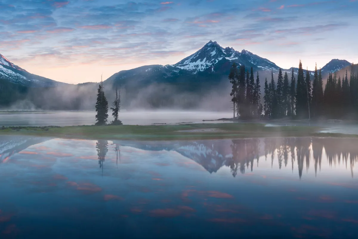 A tranquil view of mountains, a lake, and trees, with the sky reflected in the calm water. The serene landscape encourages relaxation and appreciation of the scenic surroundings.