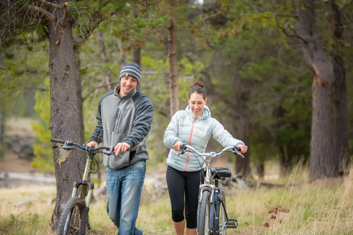 This exterior image features two cyclists walking with their bikes along a path surrounded by tall trees and lush natural landscape. The scene embodies camaraderie, outdoor activity, and the spirit of exploring nature with others.