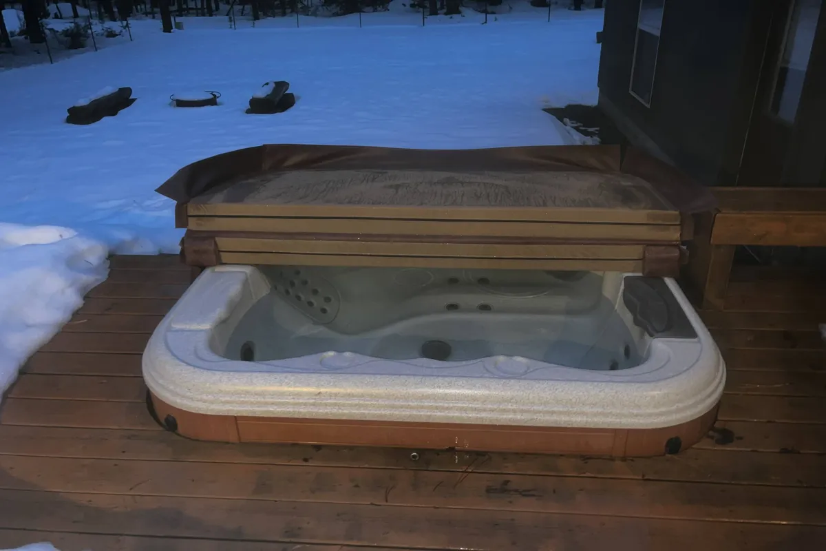 A snow-dusted wooden deck features a steaming private hot tub, with a serene backdrop of wintry landscape. The scene captures the inviting warmth of the hot tub amidst a chilly evening, hinting at year-round enjoyment and relaxation.