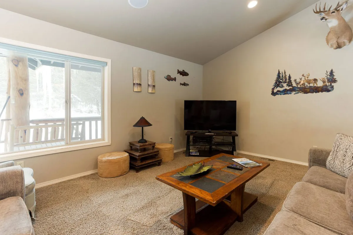 A cozy living room with a large window offering a view outside, a flat-screen TV, wooden coffee table, decorative fish art, and a mounted deer head. The space is arranged for relaxing and gathering, with ample room for family and pets to be together.
