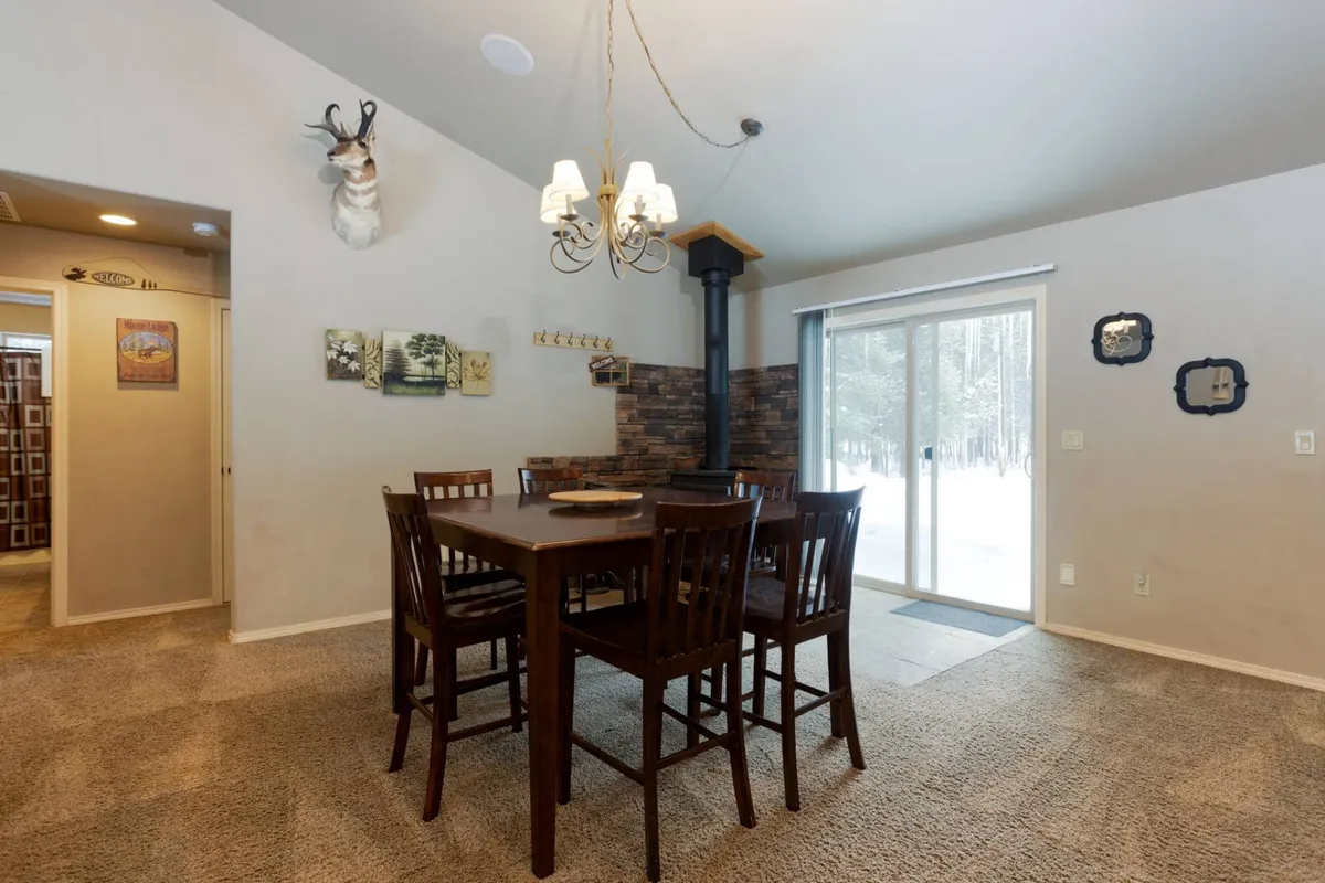 The photo highlights a rustic dining area with a large table, a wood-burning stove, and a sliding glass door that lets in natural light. The space is decorated with a mounted deer head, enhancing the warm, cabin-like feel. Ample seating makes it perfect for family meals and storytelling.